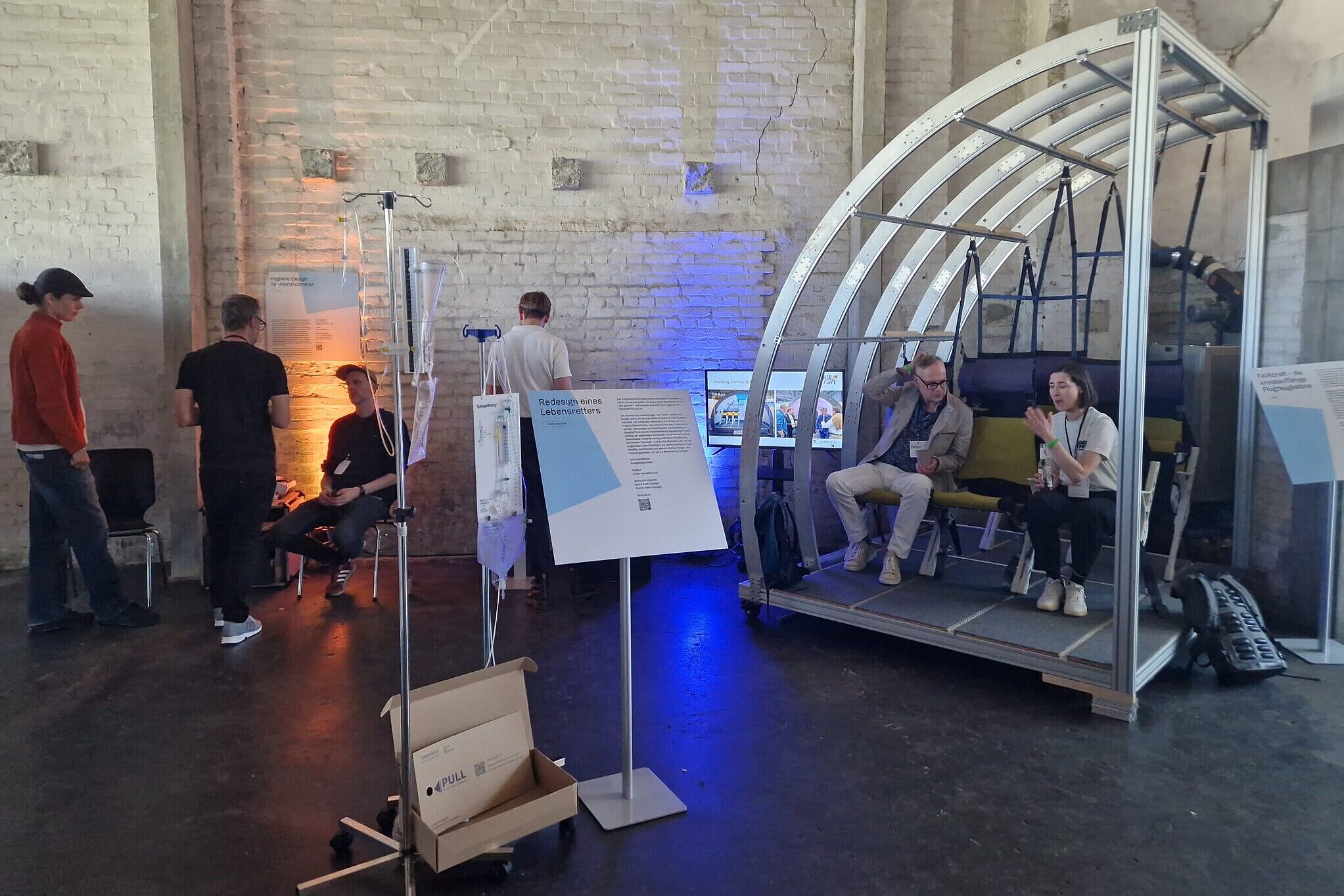 Zu sehen ist eine kühl wirkende Veranstaltungsfläche mit verschiedenen Ausstellungsstücken. Darunter ist eine große Sitzbank, die durch ihre rohe und minimalistische Metall-Überdachung wie ein Wagon wirkt. Im entfernten Hintergrund sind Menschen zu erkennen, die sich Ausstellungsstücke ansehen. 