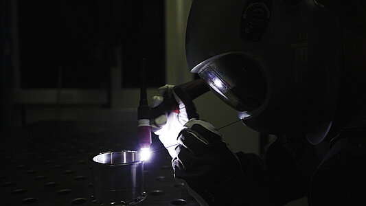 Eine Person trägt einen Schweißhelm und führt Schweißarbeiten an einem Metallstück durch.