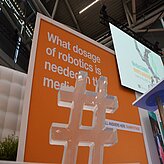 Ein großes oranges Schild mit weißer Aufschrift ist zentral im Bild zu sehen. Davor steht ein großes, transparentes Hashtag-Symbol. Im Hintergrund sind Teile einer Deckenstruktur sowie ein weiterer Bildschirm oder eine Tafel mit unscharfen Texten und Grafiken erkennbar