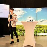 Zu sehen ist eine Referentin auf der Bühne, die Ihren Vortrag präsentiert