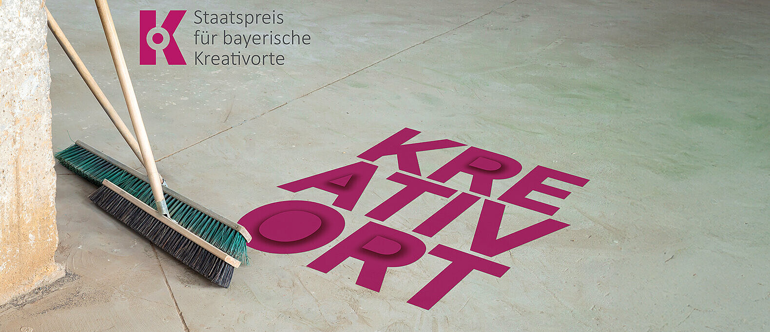 Das Bild zeigt eine glatte Betonfläche, auf der in großen, kräftigen pinken Buchstaben das Wort „KREATIVORT“ steht. Links lehnen zwei Besen an einer Wand, die Borsten berühren den Boden. Oben links ist das Logo der Auszeichnung „Staatspreis für bayerische Kreativorte“ zu sehen, bestehend aus einem stilisierten „K“ in Pink und dem Schriftzug daneben. Der Bildaufbau vermittelt den Eindruck eines vorbereiteten oder aufgeräumten Raumes, der mit Kreativität in Verbindung gebracht wird.