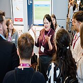 Frau mit lila Pullover und Namensschild spricht vor einer Gruppe von Menschen mit lila und roten Ausweisbändern in einem Messe- oder Konferenzraum