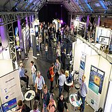 Der Blick von oben zeigt eine Ausstellung mit zahlreichen Messe-ständen. Besucherinnen und Besucher bewegen sich zwischen den Ständen und führen Gespräche.