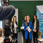 Vier Frauen stehen vor einem grünen Hintergrund und halten Papiere, im Vordergrund ist eine Kamera zu sehen