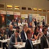 Fachbesucherinnen und -besucher folgen konzentriert dem Programm, im Hintergrund Infostände von Bayern Innovativ und Partnern.