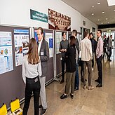Das Bild zeigt mehrere Personen, die sich in einem Flur oder Ausstellungsbereich befinden und verschiedene Poster auf Stellwänden betrachten und darüber diskutieren. Die Poster scheinen akademische oder fachliche Inhalte zu präsentieren.
