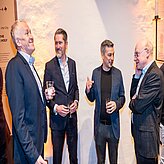 Das Bild zeigt vier Personen, die in einem Halbkreis stehen und sich miteinander unterhalten. Alle tragen formelle Kleidung, darunter Anzüge und Hemden. Der Hintergrund besteht aus einer schlichten, strukturierten Wand, auf die Licht fällt und Schatten wirft.