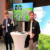 Auf der Bühne stehen an einem Stehtisch zwei Referenten, im Hintergrund ist eine Videoleinwand sowie ein Aufsteller mit dem Motiv einer Wiese und Himmel 