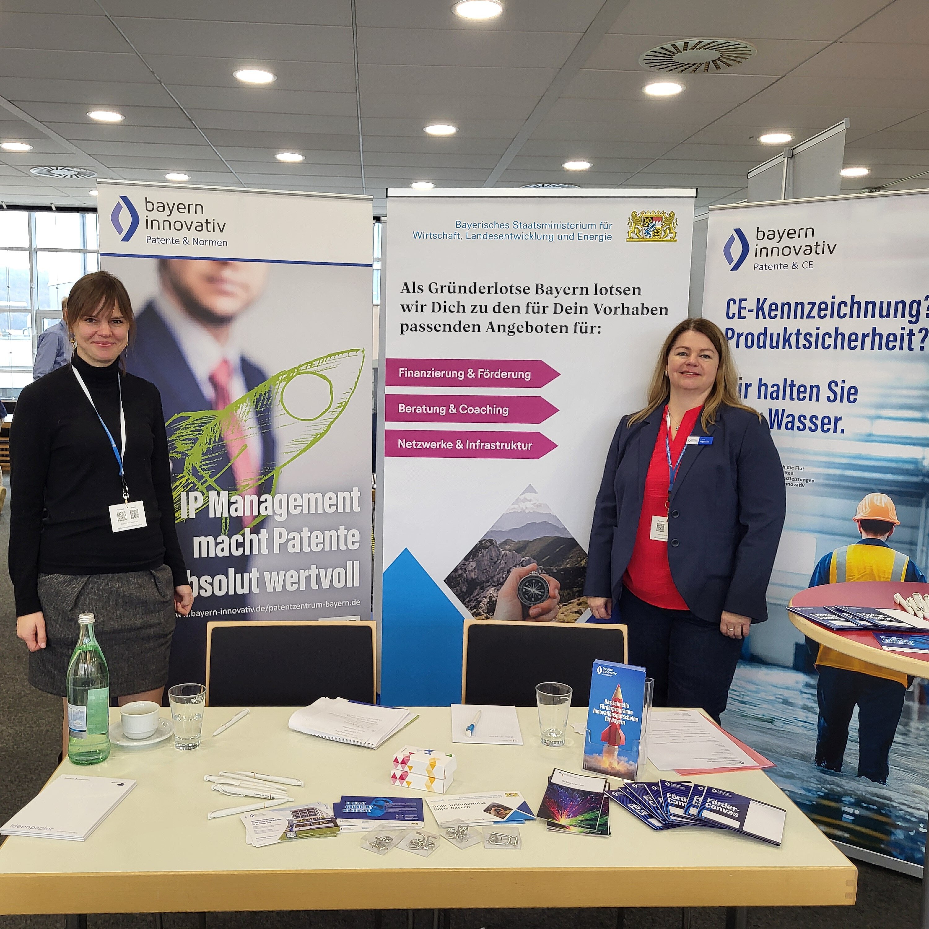 Charlotte Stegmayer, Projektmanagerin des Innovationsnetzwerk Kreativwirtschaft der Bayern Innovativ GmbH und Katrin Wägemann, Projektmanagerin Patente und CE der Bayern Innovativ GmbH, stehen hinter einem Tisch. Vor ihnen liegen ausgebreitet etliche Flyer und Stifte. Im Hintergrund stehen drei Roll-ups.