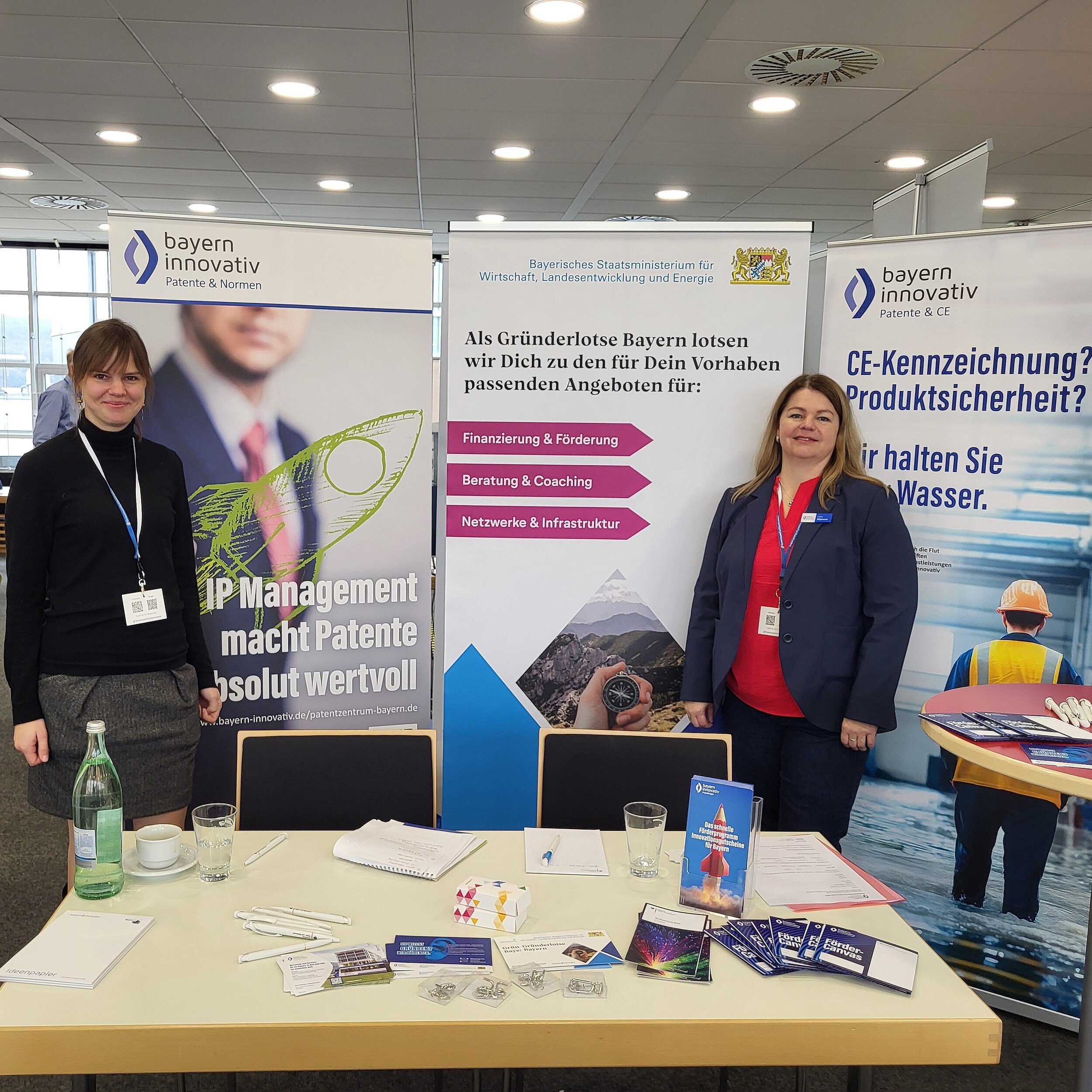 Charlotte Stegmayer, Projektmanagerin des Innovationsnetzwerk Kreativwirtschaft der Bayern Innovativ GmbH und Katrin Wägemann, Projektmanagerin Patente und CE der Bayern Innovativ GmbH, stehen hinter einem Tisch. Vor ihnen liegen ausgebreitet etliche Flyer und Stifte. Im Hintergrund stehen drei Roll-ups.