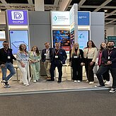 Auf dem Bild ist eine große Gruppe von Personen zu sehen, welche vor einem Messestand posieren.