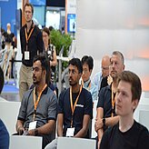 „Eine Gruppe von Männern und Frauen sitzt auf Stühlen und blickt nach vorn, als würde sie an einer Präsentation oder einem Vortrag teilnehmen. Einige Personen tragen Namensschilder an Lanyards, was auf eine Konferenz oder Veranstaltung hinweist.“