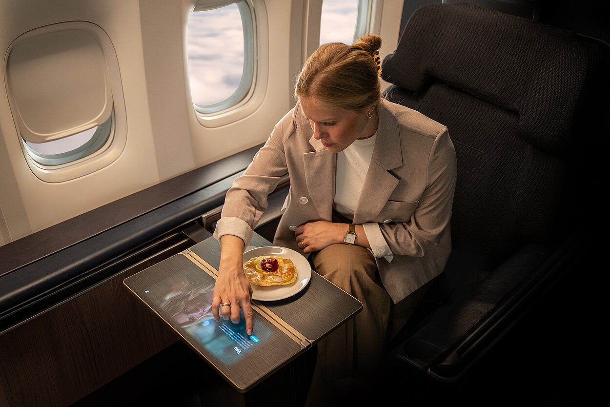 Das Bild zeigt eine Frau in einem Flugzeug, die auf einem Display tippt, welches in die Tischplatte integriert ist