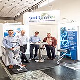 "Vier Personen stehen vor einem Messestand mit der Aufschrift 'softgate'. Links und rechts des Standes befinden sich zwei hohe Banner mit Symbolen und Text. In der Mitte steht ein kleiner runder Tisch mit Materialien darauf, umgeben von einigen Hockern. Der Boden ist mit dunklem Teppich ausgelegt."