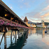 Das Bild zeigt die Kapellbrücke in Luzern mit blumengeschmücktem Holzdach über dem Wasser. Im Hintergrund sind die Jesuitenkirche, weitere Gebäude und Berge bei stimmungsvoller Abendbeleuchtung zu sehen.