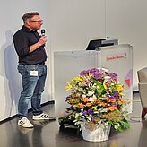 Prof. Daniel Nusser, Professor und Studiengangsleitung für Prozess- und Produktdesign, DIP-LOMA Hochschule, ist zu sehen, wie er am Pult ins Mikrofon spricht, während vor ihm eine Vase mit Blumen steht.