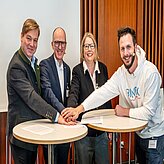 Bild zeigt vier Personen, die um zwei kleine runde Tische stehen und ihre Hände in der Mitte zusammenlegen