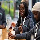 Das Bild zeigt eine junge Frau mit Brille und Zöpfen, die an einem Tisch sitzt und sich angeregt unterhält. Sie trägt einen grauen Hoodie mit dem Aufdruck "HEALTHCARE HACKATHON BAYERN MÜNCHEN". Neben ihr sitzt ein Mann mit Mütze, der ebenfalls einen Hoodie mit dem gleichen Aufdruck trägt.