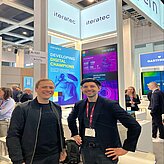 Auf dem Bild sind zwei in schwarz gekleidete Herren zu sehen, welche vor einem Messestand posieren.