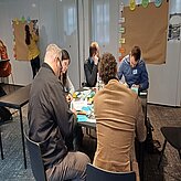 Auf diesem Bild ist eine Gruppe von Menschen zu sehen, die um einen Tisch sitzen und sich mit einer Aktivität oder Diskussion beschäftigen.