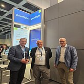 Auf dem Bild sind drei Herren im Anzug zu sehen, welche vor einem Messestand posieren.