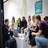 Das Bild zeigt junge Erwachsene bei einer informellen Besprechung während eines Hackathons. Im Hintergrund hängt ein Poster für den "Healthcare Hackathon Bayern". Die Atmosphäre ist konzentriert und kollaborativ.