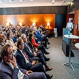 das Bild zeigt eine Konferenz- oder Seminarveranstaltung. Eine Person steht vorne im Raum und spricht zu einem Publikum, das in Reihen sitzt und aufmerksam zuhört. Der Raum ist mit Holzvertäfelung gestaltet und durch Lampen an der Rückwand gut beleuchtet.