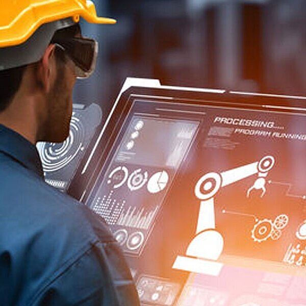 Ein Arbeiter mit Schutzhelm und Schutzbrille steht vor einem großen Display in einer Produktionshalle