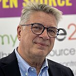 Auf dem Bild ist ein Herr mit grauen Haaren und Brille zu sehen. Er trägt ein hellblaues Hemd und ein dunkles Jacket und lächelt in die Kamera.