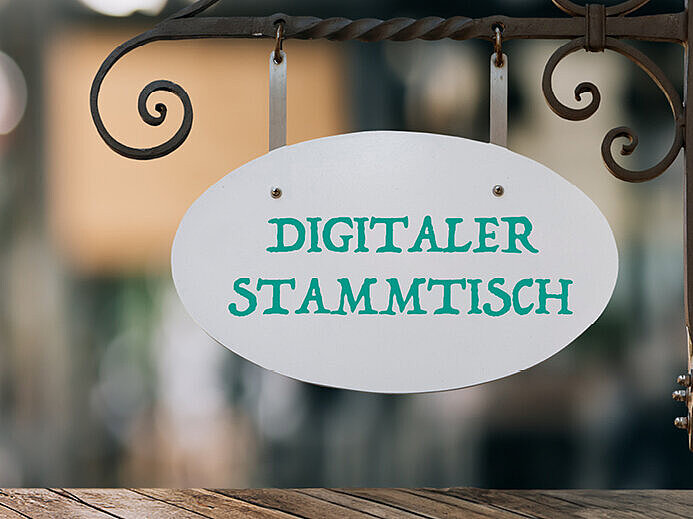 Auf dem Bild ist ein Banner für den "Digitalen Partnerstammtisch Gesundheit". Im Hintergrund sind die verschwommenen Details eines Biergartens zu sehen.