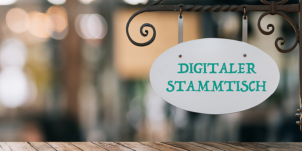 Auf dem Bild ist ein Banner für den "Digitalen Partnerstammtisch Gesundheit". Im Hintergrund sind die verschwommenen Details eines Biergartens zu sehen.