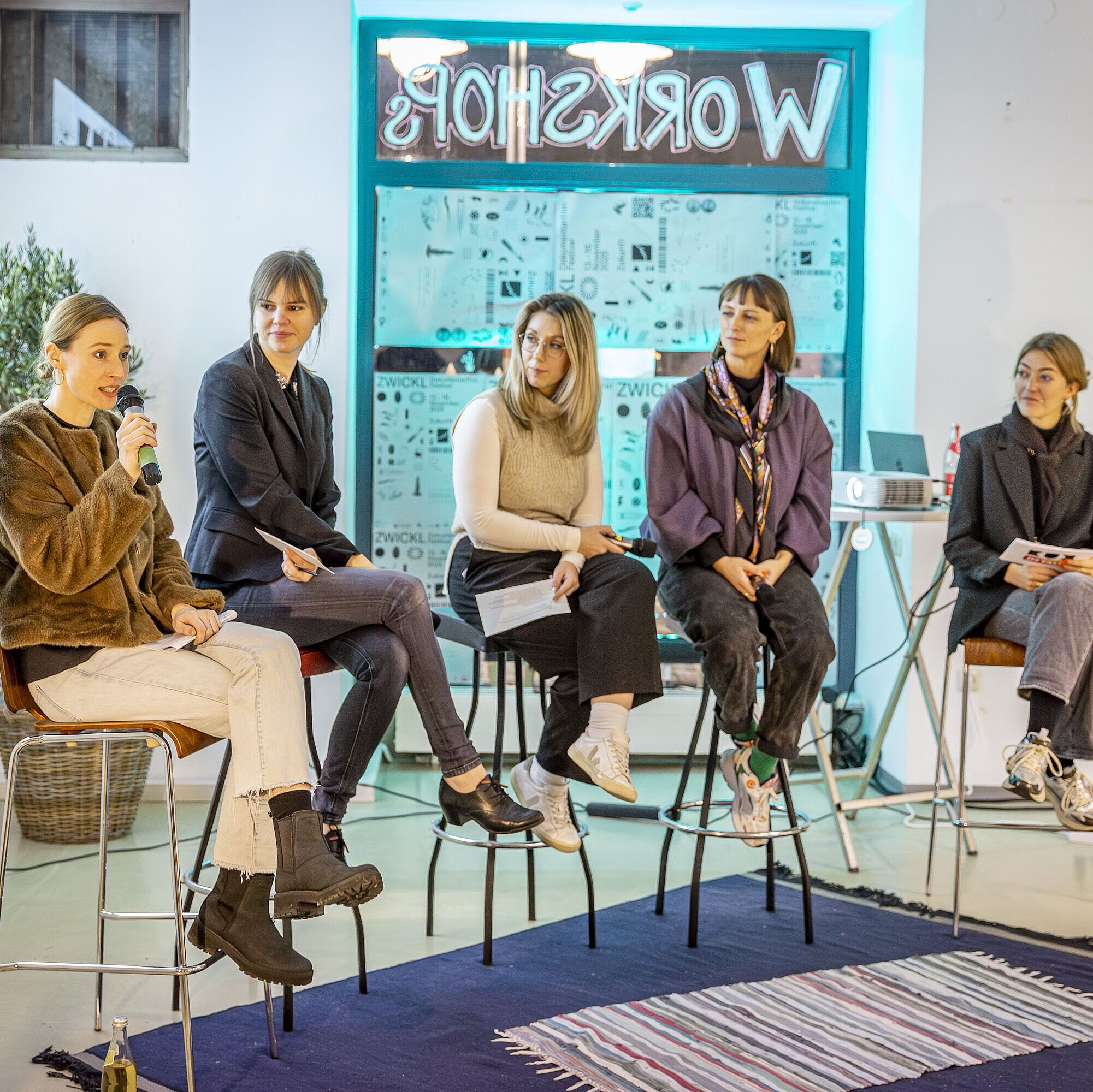 Auf dem Bild sitzt ein fünfköpfiges Panel, darunter Charlotte Stegmayer, Projektmanagerin im Innovationsnetzwerk Kreativwirtschaft der Bayern Innovativ GmbH, auf hohen Hockern vor einem Publikum. Alle halten Mikrofone oder Notizen in der Hand. Im Hintergrund sind mehrere Poster zu sehen.