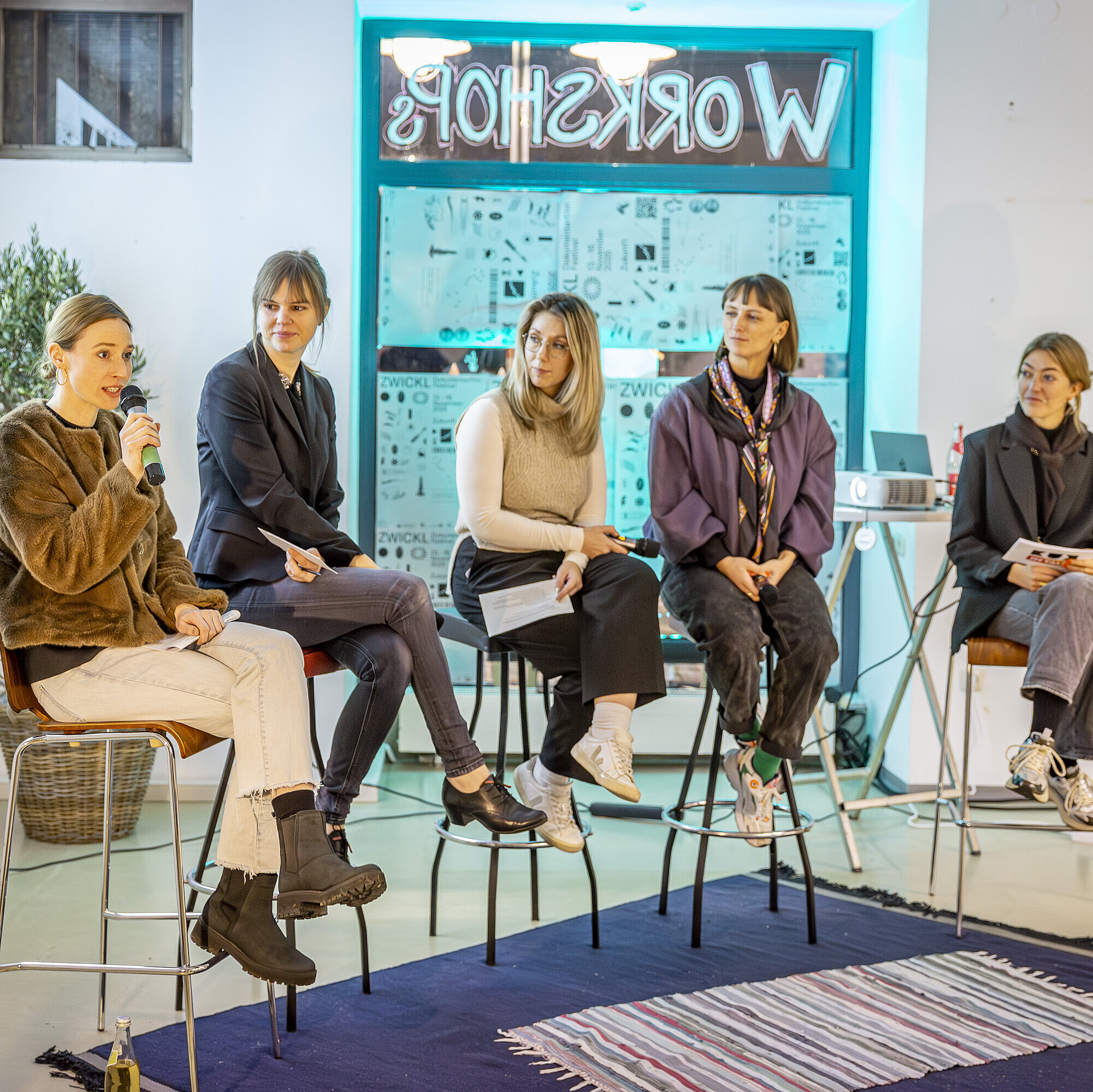 Auf dem Bild sitzt ein fünfköpfiges Panel, darunter Charlotte Stegmayer, Projektmanagerin im Innovationsnetzwerk Kreativwirtschaft der Bayern Innovativ GmbH, auf hohen Hockern vor einem Publikum. Alle halten Mikrofone oder Notizen in der Hand. Im Hintergrund sind mehrere Poster zu sehen.