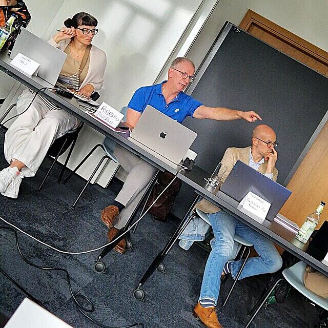 Blick auf vier Personen, die an einem Workshoptisch sitzen. Die eine männliche Person zeigt  nach Rechts.