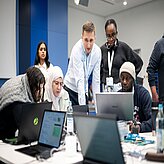 Menschen sitzen an Laptops und arbeiten, im Hintergrund stehen zwei Männer und beobachten