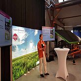 Ein Referent auf der Bühne mit Stehtisch und im Hintergrund eine Motivwand auf der ein Acker mit weißblauem Himmel abgebildet ist