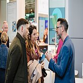 Drei Personen im Gespräch an einem Messestand, eine Person hält eine Tasse, zwei Personen tragen Mäntel und halten Broschüren