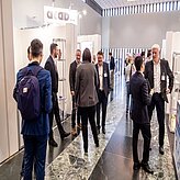 das Bild zeigt eine Gruppe von Personen, die sich in einem Innenraum – vermutlich im Rahmen einer Konferenz oder Veranstaltung – unterhalten. Mehrere Personen tragen Businesskleidung, einige halten Unterlagen oder Mappen in der Hand