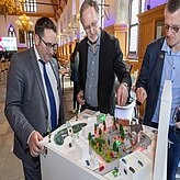 Drei Männer betrachten ein Stadtmodell, das auf einem Tisch präsentiert wird, während sie angeregt diskutieren.