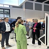 Auf dem Bild sind drei Personen in einem Veranstaltungsraum zu sehen. Eine Frau ist mit dem Rücken zugewandt und trägt einen grünen Hosenanzug.