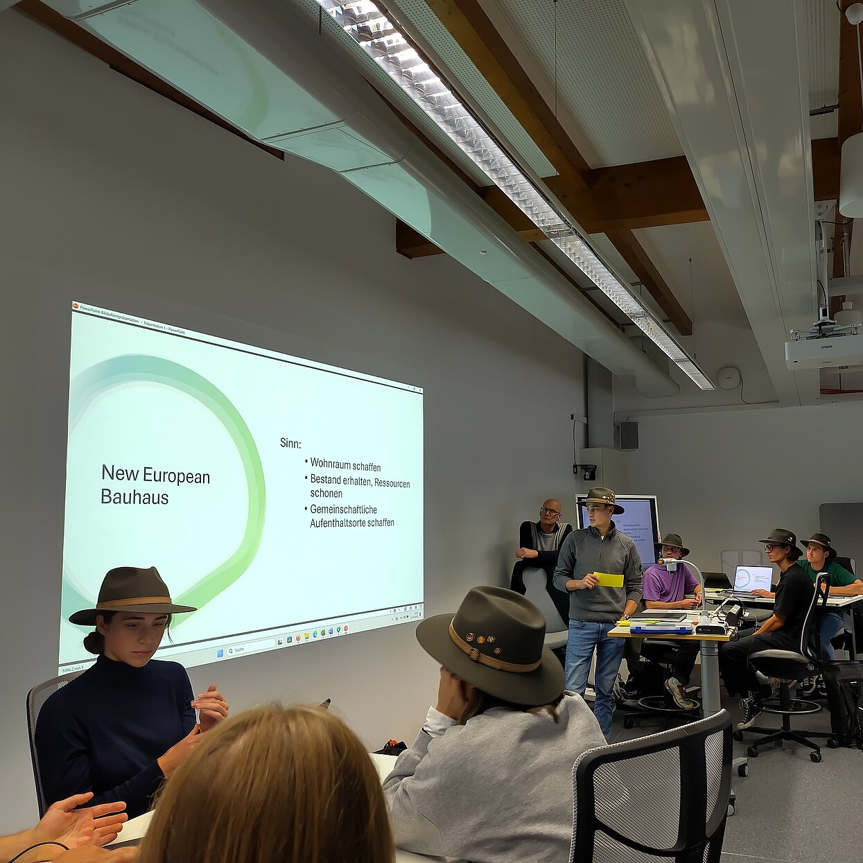 Eine Präsentation mit New European Bauhaus. Davor sitzen Studierende mit un dohne grünen Hüten.
