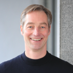 Mann mit kurzem hellbraunem Haar trägt schwarzen Pullover und steht vor einer grauen Wand