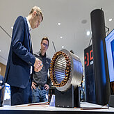 Zwei Männer stehen neben einem Fahrzeugteil, dass im Rahmen der Ausstellung präsentiert wird. 