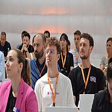 „Eine Gruppe von Personen steht eng beieinander und hält Blätter oder Karten in die Kamera. Der Fokus liegt auf den Gesichtern der Personen und den hochgehaltenen Papieren.