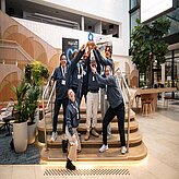 „Gruppen von Teilnehmenden sitzen an Tischen und arbeiten konzentriert an Laptops während eines Hackathons, im Hintergrund sind Präsentationsflächen und Banner sichtbar.“