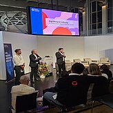 Begrüßung & Eröffnung: Robert Bartl, Mitglied der Geschäftsleitung, Bayern Innovativ GmbH, spricht vor Publikum in ein Mikrofon, während Markus Eder, Ministerialdirigent, Bayerisches Staatsministerium für Wirtschaft, Landesentwicklung und Energie und Christian Fayek, Leitung Projekte, bayern design GmbH, neben ihm stehen.