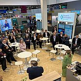 Auf dem Bild ist ein Veranstaltungsraum mit Stehtischen und Messeständen zu sehen, in welchem sich ein paar Personen befinden und sich einen Vortrag anhören.