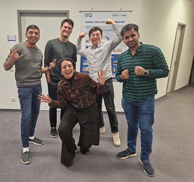  Fünf Personen posieren zusammen in einem Raum. Vier von ihnen stehen, und eine Person kniet vorne. Sie machen verschiedene Handgesten und scheinen Spaß zu haben. Im Hintergrund sind eine Tür, eine Wand mit einigen Postern oder Schildern und eine offene Tür zu einem anderen Raum zu sehen.