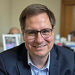 Auf dem Bild ist ein Herr mit braunen Haaren und Brille zu sehen. Er trägt ein hellblaues Hemd und ein dunkles Jacket und lächelt freundlich in die Kamera.