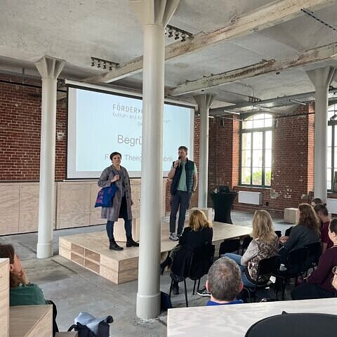 In einem Vortragsraum stehen zwei Personen vor einer Präsentation auf der Bühne vor einem befüllten Raum. 